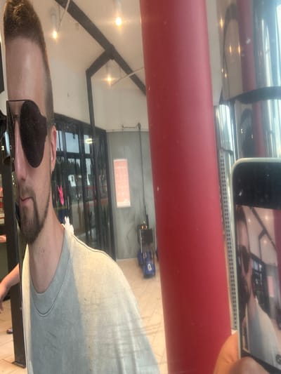 The image is a low-angle selfie taken by a person wearing sunglasses and a light gray t-shirt. The person has a goatee and short, dark hair. They are standing in what appears to be an indoor public space with a tiled floor and a ceiling with visible lighting fixtures and dark beams. To the right of the person is a large, bright red cylindrical pillar. A smartphone is held up, capturing a reflection of the person in its screen, as well as a distorted view of the surroundings. In the background, through glass doors, there are reflections of trees and outdoor elements, suggesting the location is near an entrance or exit. A pink decoration is visible hanging from the doorway in the background. There are also some shopping carts or trolleys visible in the mid-ground to the right. The overall lighting is bright, and the image has a slightly overexposed quality. The location is not specified as Slitu, Norway.