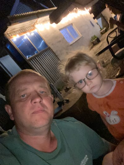 This is a selfie-style video taken outdoors at night, with a focus on a man and a young child. The man, positioned in the foreground and slightly to the right, is looking towards the camera with a relaxed expression. He is wearing a green t-shirt with a logo on the left side of his chest that appears to read "science...". The child, who is to the left and slightly behind the man, is wearing glasses and an orange outfit with a white bunny applique on the chest. The child is looking away from the camera, seemingly at something out of frame.
The background shows a building with white walls and windows, some of which are lit from within, suggesting it's an interior space. A string of lights, likely fairy lights or cafe lights, is draped along the building, casting a warm glow. The lighting suggests it is nighttime. The overall scene appears to be a residential or possibly a recreational outdoor area, given the presence of the building and decorative lighting. The orientation of the video is such that the subjects are nearly upside down. There are no discernible weather cues, but the lighting implies it is not daylight. The context of "Әуезов ауылдық округі, Kazakhstan" places the scene in a rural or semi-rural area of Kazakhstan.