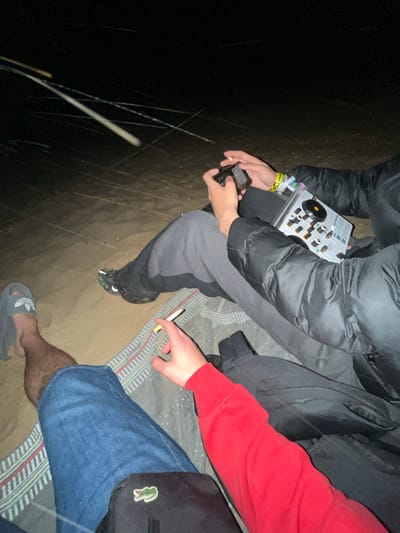 The image shows two people sitting on a blanket outdoors at night. One person, wearing a black puffer jacket and grey sweatpants with black shoes, holds a small electronic device (likely a vape or portable music player) in one hand and a DJ controller in their lap. The other person, wearing a red long-sleeved shirt and blue denim shorts with grey slide sandals, holds a lit cigarette. A black shoulder bag with a Lacoste logo is visible near them. The setting appears to be dark, possibly on a beach or other outdoor area with sand, inferred from the ground texture. Given the request for context, this scene could be near a beach in Punta Umbría, Spain.