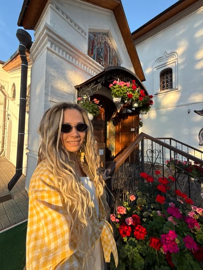 A young woman with long blonde dreadlocks and dark oval sunglasses stands smiling in front of the Krestovozdvizhensky Khram (Exaltation of the Holy Cross Church) in Kuznechikha, Russia. She wears a yellow and white gingham long-sleeved shirt over a white top. The church, constructed in 1757, features white stucco walls with classical architectural details. Its brown wooden main entrance is framed by a black ornate metal canopy, from which multiple white hanging baskets filled with red and pink geraniums are suspended. Above the entrance, an arched niche contains a vibrant mosaic depicting religious figures. To the left of the entrance, a white plaque with Russian text details the church's name and construction year. A black downspout with silver bands is visible on the far left. A set of brown wooden stairs with black wrought-iron railings, adorned with heart-shaped motifs, ascends to the entrance, flanked by numerous planters brimming with red and pink geraniums. To the right of the entrance, a small, arched window with a decorative metal grille is set into the white wall. Warm, low-angle sunlight illuminates the scene, creating shadows and highlighting the building's facade and the vibrant flowers. A security camera is visible on the wall to the right of the entrance. The ground to the left is paved with light-colored blocks, with a patch of green matting in the foreground.