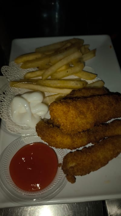 This forensic description details a meal served on a white rectangular plate, located in Sena, Indonesia. The plate holds multiple golden-brown, breaded and fried chicken strips and a mound of golden-yellow french fries. A small portion of white mayonnaise is presented on a white doily adjacent to the fries. Separately, a clear plastic cup contains a red sauce, likely ketchup or chili sauce. The background is dark and out of focus, indicating an indoor setting. A metallic, reflective surface occupies the lower edge of the frame. No individuals, actions, or interactions are visible; the scene depicts a static food arrangement.