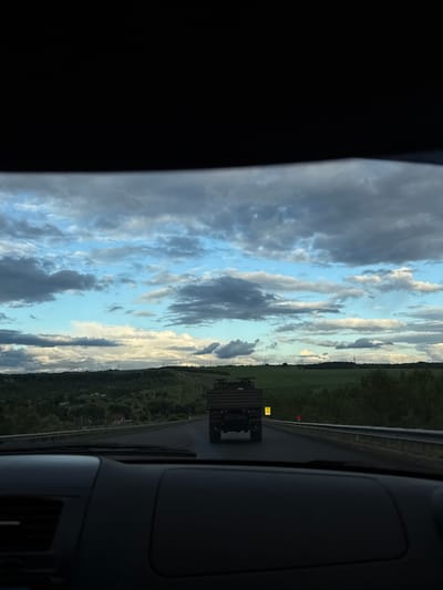 The image is taken from inside a vehicle, looking out through the windshield. The scene is outdoors, with a road stretching into the distance and a cloudy sky above. In the foreground, the dashboard of the vehicle is visible, with a dark, textured surface. Ahead on the road, a military truck is driving away from the camera. The truck is olive drab and has a flatbed with a raised, slatted rear section. The road is bordered by a guardrail and then by green, hilly terrain covered in trees and fields. Power lines are visible on the horizon. The sky is filled with a mix of blue sky and dramatic grey and white clouds. There are no people visible in the image. The overall impression is of a scenic drive through a rural landscape.