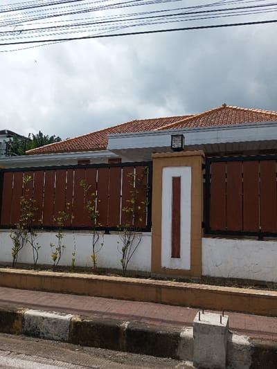 This image depicts a residential scene in Viengthong, Laos, under an overcast sky with overhead power lines. A modern house with a reddish-orange tiled roof and white fascia is partially visible behind a perimeter fence. The fence features dark brown vertical slats set in black frames, supported by a lower white concrete wall exhibiting cracks and discoloration. Small, leafy green plants are cultivated along the base of the fence. A prominent tan and white concrete pillar, adorned with a recessed dark brown vertical stripe and topped with a black rectangular lantern, forms part of the fence structure. In the foreground, a reddish-brown tiled sidewalk is bordered by a concrete curb with alternating black and white sections, showing wear and grime. A small, unpainted concrete post with four exposed rebar rods protrudes from the sidewalk's edge on the right. A dark road surface is partially visible at the bottom. A small, faint white object, possibly an insect or bird, is visible against the sky near the upper left. No individuals or active interactions are present.