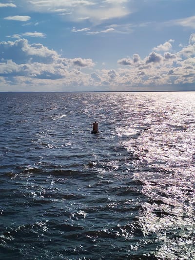 A view of a wide, dark blue body of water under a bright, partly cloudy sky. The sun is shining, creating a glittering reflection on the water's surface, with a distinct path of light stretching towards the horizon. A small, red-brown buoy with a dark cap sits in the water, slightly to the right of the center. The horizon line is visible, separating the water from the sky, with a faint, hazy landmass in the distance. The sky is a mix of blue with scattered white, fluffy clouds, some of which are catching the sunlight. The overall impression is one of a peaceful, sunny day on a large body of water, possibly a lake or sea. The location is identified as Myaksa, Russia. The time of day appears to be daytime, likely mid-morning or afternoon, given the bright sunlight and clear sky. There is no visible text in the image.