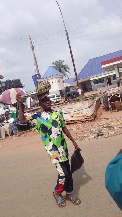 An individual, appearing male, is captured walking on an asphalt road under an overcast sky in Igbo Ukwu, Nigeria. The person is wearing a green short-sleeved t-shirt patterned with Mickey Mouse and Donald Duck characters, dark blue track pants with white and red vertical stripes, and grey Nike slide sandals. A silver-colored flat tray, holding multiple items wrapped in green leaves, is balanced on their head. Their right hand is raised, positioned near the tray, while their left hand holds a black plastic bag at waist level. The individual's mouth is open. The background features various structures, including buildings with prominent blue roofs, one of which has a red canopy, suggesting a commercial establishment like a fuel station. A wooden utility pole with wires, a reddish-brown street light pole, a palm tree, a white van, and a tricycle are also visible. Areas off the road contain unpaved ground with debris and rudimentary market stalls, one covered by a purple and white striped umbrella.