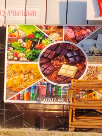 The image shows a colorful collage of food items displayed on what appears to be a storefront or market stall. The collage is divided into several sections, each featuring a different type of food.

In the top left, there's a depiction of fresh produce, including a basket overflowing with vegetables like broccoli, carrots, tomatoes, and cauliflower. To its right, a section shows various types of cured meats, some with red and white checkered wrapping, suggesting a deli or charcuterie selection.

The bottom left displays a variety of pasta shapes and possibly some fried snacks. In the center, a large circular section showcases an assortment of chocolate bars, with a prominent piece of white chocolate on top of darker chocolate pieces. Adjacent to this, there are images of what seem to be nuts or candy.

At the bottom of the collage, a row of colorful canned beverages, likely sodas, are visible. Below the collage, there is a phone number displayed: "0707 49 19". The background behind the collage appears to be a white wall with some vertical dividers. To the right of the collage, a wooden shelf unit is visible, with some items on display. The overall impression is that of a food vendor's advertisement. The image is well-lit, suggesting it was taken during the day. There are no people visible in the image. The scene is likely an outdoor or semi-outdoor market setting. The text "ИЗ АЧЫЛДЫК" is visible on a white sign above the collage, which might be a store name or a greeting. Given the location context of Ala-Buka, Kyrgyzstan, the text could be in Kyrgyz or Russian.