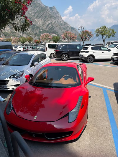 A red Ferrari sports car is parked in a designated parking spot, marked by blue lines, in Limone sul Garda, Italy. The car's interior is visible, featuring tan leather seats and a steering wheel. In the background, a white hatchback and other vehicles are parked. Beyond the cars, a picturesque view of Lake Garda is visible, with mountains rising in the distance and trees with pink blossoms adding to the scenery. A cluster of red flowers and green leaves are in the foreground. The sky is clear with scattered clouds.