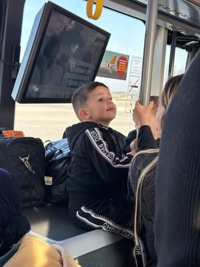 The scene depicts the interior of an airport shuttle bus, positioned on an airfield tarmac at Sabiha Gökçen International Airport (SAW) in Pendik, Istanbul, Turkey, a major transportation hub for the adjacent district of Tuzla.

In the foreground, a young male child, approximately 4-6 years old, is seated on a dark surface, possibly a bench or luggage area, looking upwards and slightly to the right with a soft, open-mouthed expression. He has short, dark hair and is wearing a black tracksuit featuring a white and black striped pattern with "CHRISTIAN DIOR" lettering along the sleeves and lower pant legs. To the child's left, multiple pieces of dark luggage are stacked, including a black checkered rolling suitcase with an orange "CABIN & MAIN BAGGAGE" tag, and a black duffel bag.

On the right side of the frame, partially obscured, is an adult individual, likely female, whose blonde hair and a hand gripping a vertical metal support pole are visible. This suggests the adult is either standing or steadying themselves within the moving vehicle. Reflections of other passengers are faintly visible on the window and an overhead screen.

An angularly mounted rectangular display screen is positioned above the child, reflecting interior elements and displaying Turkish text that translates to "Order during flight, enjoy your journey." Adjacent to the screen, visible through the window, are several informational decals: a red, heart-shaped advertisement featuring a white airplane and "PEGASUS" branding, an "EMERGENCY EXIT" sign in English with instructions to "break the window," and a standard 'no smoking' symbol.

Outside the window, a brightly lit airfield tarmac is visible under a clear sky. Distant airport infrastructure and what appears to be the tail or wing of an aircraft can be discerned, consistent with an active airport environment. Yellow grab handles are visible along the bus ceiling.