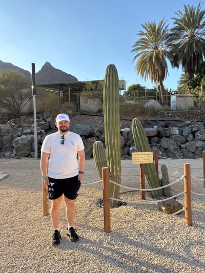 A full-body photograph captures one adult male standing on a light gravel surface in Hatta, United Arab Emirates. The subject is a light-skinned male with a short beard, wearing a white baseball cap with a light blue logo, a white short-sleeved t-shirt with a light blue palm tree graphic, black sunglasses tucked into his collar, black shorts with white graphics including "DR" and a bull logo, black athletic shoes, and white ankle socks. A blue smartwatch is on his left wrist, and a black watch is on his right. He stands facing the camera, with his hands at his sides.
To the subject's right, three large columnar cacti are visible, with the tallest extending beyond the top frame. A brown wooden sign with white lettering identifies one of the cacti as "Pachycereus Pringlei" in English and Arabic. The cacti are enclosed by a low barrier consisting of wooden posts connected by a thin rope.
In the background, under a clear blue sky, a rugged, rocky mountain peak is visible on the left. Below the mountain, sparse dry vegetation, a building with a metal roof, and a chain-link fence are present. A tall, dark pole stands vertically in the mid-ground. To the right, several mature date palm trees are visible against the sky, along with more scrub brush and additional distant structures. The ground is consistently covered in light-colored gravel, and distinct shadows indicate clear, direct sunlight, suggesting either morning or late afternoon.