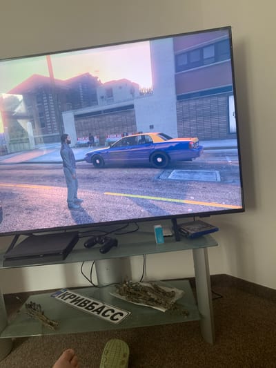 The image shows a television screen displaying a scene from a video game, likely Grand Theft Auto V, given the graphics and vehicle. The game depicts a male character standing on a street next to a purple and yellow taxi. The character is wearing a grey vest over a blue shirt and grey pants, with grey shoes. Behind the character and taxi, buildings with windows and advertisements are visible.
The television is placed on a glass and metal media console. On the console are a black gaming console, a black game controller, a small blue and white box, and a video game case. To the right of the console, on the lower shelf of the media console, is a white license plate with black Cyrillic lettering that appears to read "КРИВБАСС" (Kryvyi Rih, an industrial city in Ukraine). There are also some dried plant materials on the shelf. The floor is carpeted.
The image itself is a photograph taken from an angle, showing the television screen and the media console below it. The lighting in the room appears to be dim, with light emanating from the television screen illuminating the foreground. There is no indication of the location being Vejprty, Czechia; this detail is not supported by any visual evidence in the image.