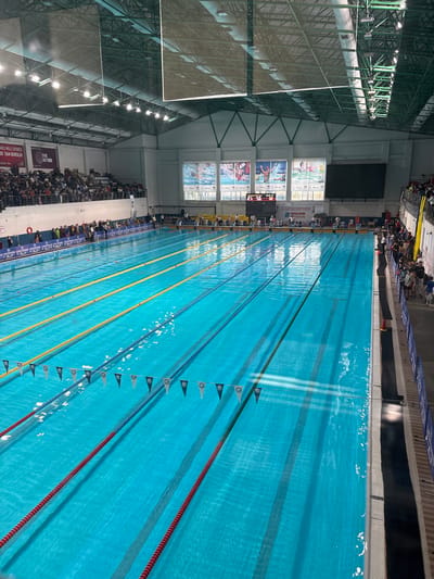 The image shows a long, indoor swimming pool with multiple lanes marked by colorful dividers. Spectators sit on both sides of the pool. The pool is located in Gungoren, Turkey, according to a sign visible in the image. Overhead lighting illuminates the pool area. A large screen and other signage are visible on the left side of the frame. There are people sitting on both sides of the swimming pool.