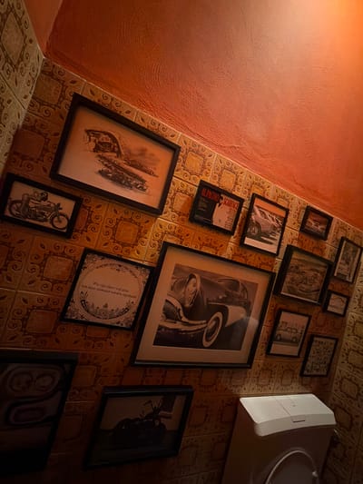 The image shows a wall with a collection of framed pictures and tiled surfaces. The tiles have a brown and beige patterned design. Above the tiled area, the wall is painted a textured orange color, with a bright light source illuminating the upper right portion of the scene. The framed pictures are predominantly black and white, and depict various subjects including vintage cars, motorcycles, and a poster for "Scarface" featuring Al Pacino. One frame contains a circular design with text in what appears to be Dutch. A white waste bin is visible in the lower right corner. The overall impression is of a decorated wall, possibly in a bathroom or a themed room, located in Varsseveld, Netherlands.