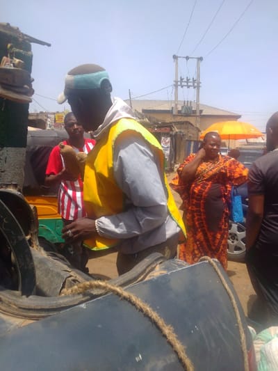 This daytime image depicts an active street scene in Mista Ali, Nigeria, characterized by bright sunlight and visible utility infrastructure. In the immediate foreground, a dark, possibly metallic, surface of a vehicle or container is visible, secured by a thick, natural fiber rope.
Centrally positioned, with his back largely to the viewer, is a male individual wearing a light grey long-sleeved shirt underneath a bright yellow safety vest featuring reflective trim. He wears a multi-colored cap (light blue, white, tan) backward. His right hand, adorned with a ring on the middle finger, is positioned near the foreground object.
Behind him and slightly to the left, a second male stands, facing forward, wearing a red and white vertically striped shirt. He holds two brown, elongated, root-like objects in his left hand. Adjacent to him is the yellow and green bodywork of a motorized tricycle (Keke Napep).
To the right of the central figure, a woman stands, facing slightly left, wearing a vibrant orange and black patterned dress. She carries a bright blue reusable bag in her left hand and has her right hand near her ear. A partial view of another individual in a black shirt is visible on the far right.
The background reveals a typical urban environment with various single-story buildings and market stalls lining an unpaved, dusty street. Overhead, a utility pole with a transformer and multiple power lines are prominent. An open orange market umbrella provides shade on the right side of the background.