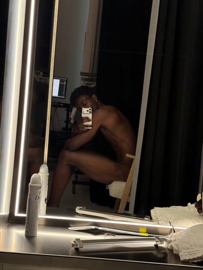 The image is a reflection in a mirror, showing a person taking a selfie with a smartphone. The person is partially undressed and is in a crouching or kneeling position. Their skin tone is dark. A wooden object is visible behind the person, and some white fabric is draped over it. In the reflection, a computer monitor is visible in the background. On the surface below the person's reflection, two spray cans are present, one of which has visible text: "Dove invisible". The lighting is artificial and bright, suggesting an indoor setting. The overall mood is intimate and personal due to the nature of the selfie and the partially undressed subject. The location context of "Fibracolor, Spain" is not directly evident from visual cues within the image itself, but it is provided as external information.