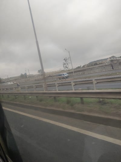 The image displays a multi-lane highway in Gatongora ward, Kenya, observed from within a moving vehicle under an overcast sky. In the immediate foreground, a dark asphalt road surface with a white lane marking is visible, bordered by a metal guardrail extending horizontally. Beyond the guardrail, a strip of green grass and dirt separates it from the further road sections. The mid-ground encompasses several asphalt road lanes, delineated by white dashed lines and separated by concrete barriers. A motorcycle (boda-boda) with two occupants is visible in one lane, moving towards the left of the frame. Further right, on a slightly elevated road section, a light-colored van is present near several small, white-and-black striped bollards. Two human figures are discernible on foot near the van, one positioned to its right and walking further right. Multiple tall utility poles, including a prominent central one and a streetlight with a curved arm, are spaced along the roadside. The background features an elevated terrain supported by a retaining wall, upon which various light-colored buildings with dark roofs are constructed, consistent with residential or semi-urban structures. The sky is uniformly gray and heavily overcast.