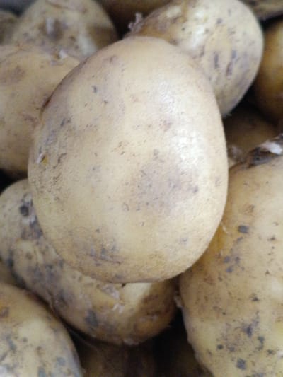 A close-up, overhead shot shows a pile of potatoes. The potatoes are light brown with some dark spots and blemishes. The potatoes are of various sizes and shapes, and they are piled on top of each other. The background is dark and out of focus. This image was taken in Martuni, Armenia.