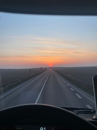 The image displays a forensic view from inside a vehicle, likely a truck, looking forward through the windshield at a two-lane road during sunset. The location is Slaný, Czechia. The sky, from a light blue overhead, transitions to vibrant orange, pink, and red hues at the horizon, where a distinct red-orange sun is visible centrally. Minimal, wispy clouds are present in the middle sky. The paved road stretches directly into the distance, exhibiting a slight curve to the right, and is delineated by solid white lines at its edges and a dashed white line separating the two directions of travel. Metallic guardrails flank both sides of the road, receding into the atmospheric haze. Flat, dark-toned fields, consistent with agricultural land, extend to the horizon on either side of the roadway. Distant, indistinct structures are faintly visible along the horizon line. The top portion of a black steering wheel is visible in the lower foreground, and a digital display beneath it shows "81," likely indicating speed, along with a "9°C" temperature icon and a green signal indicator. No individuals or other vehicles are distinctly visible within the immediate scene.