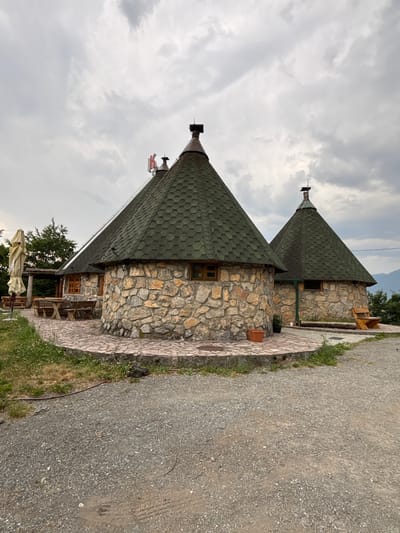 The image showcases two rustic, stone buildings with conical, green-tiled roofs, reminiscent of traditional mountain architecture. These structures are situated in a natural setting, likely in a rural or mountainous area, as suggested by the presence of trees and a cloudy sky. A gravel path runs alongside the buildings, with some grass and a few potted plants adorning the area.
The buildings themselves are constructed with rough-hewn stones, giving them a sturdy and weathered appearance. They feature small, wooden-framed windows and what appear to be chimneys or decorative elements on their pointed roofs. The roofs are covered in hexagonal tiles, creating a textured, scale-like pattern.
The scene suggests a tranquil, possibly remote location. The cloudy sky indicates overcast weather, and the lighting implies it could be daytime, perhaps late afternoon or evening. The presence of outdoor seating, including a table and chairs visible through one of the building's openings, suggests that the buildings might be part of a restaurant, guesthouse, or a similar establishment offering hospitality.
There is no visible text in the image. The overall impression is one of rustic charm and a peaceful, natural environment. The style of the buildings and the surrounding landscape align with the context of Mekića Kula, Montenegro, which is known for its scenic mountain vistas and traditional structures.