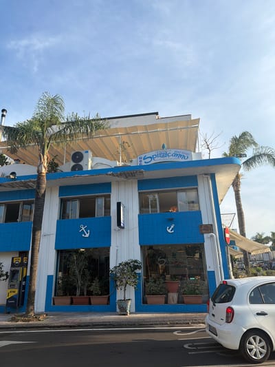 The image shows a two-story building with a blue and white facade in the city of Chianchitta, Italy. The building houses a restaurant/pizzeria called "Spizzicann." The building has blue tile accents with white anchor designs. There are windows on both stories, some with potted plants inside. An ATM is located on the ground floor. A white car is parked nearby. Palm trees are present. The sky is visible. The image is rotated 90 degrees.