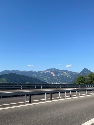 The image presents a static, elevated view of an asphalt highway in the vicinity of Buochs, Switzerland, under a clear, sunlit sky. In the immediate foreground, dark asphalt pavement is marked by two parallel, solid white lines. A grey metal guardrail, consisting of horizontal beams supported by vertical posts, extends horizontally across the lower half of the frame, serving as a barrier between the roadway and a drop-off or adjacent area. Beyond this, a secondary, darker grey concrete barrier with a lighter top rail is visible.
The midground and background are dominated by multiple ranges of mountains. These mountains are predominantly covered in dense green foliage and trees, with some higher peaks exhibiting visible light-colored rock faces. A small cluster of structures, indicative of a human settlement, is discernible at the base of the mountains in the distant left. The upper half of the frame displays an unobstructed expanse of bright blue sky, featuring two small, distinct white cumulus clouds and a single, minute dark object located in the upper left quadrant. The scene is devoid of human figures or active vehicles.