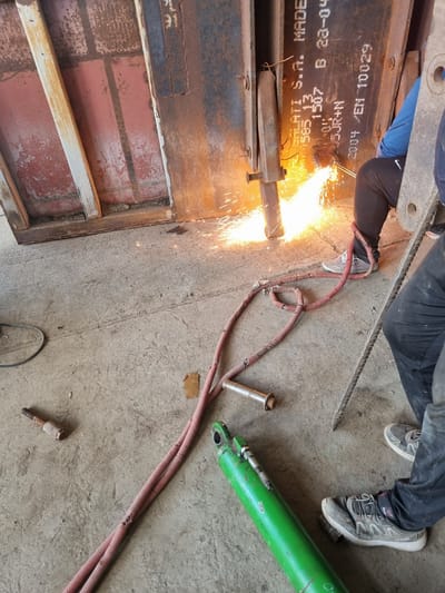 An overhead shot depicts a worker torch-cutting a metal structure, generating sparks. The worker is wearing dark pants, a blue shirt, and grey athletic shoes. A twisted, red hose stretches from the torch to a green hydraulic cylinder resting on the concrete floor. Debris is scattered around the workspace. Text "MELATI S.A. MADE" is visible on the structure. The location is unidentified, with no visible clues indicating Ovcharovo, Bulgaria.