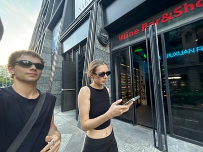 The image, oriented correctly, depicts two people, a woman and a man, standing outside a modern building with glass windows in an urban setting during daylight.

The woman, positioned on the left, has blonde hair and wears sunglasses. She is dressed in a black sleeveless crop top, revealing her midriff, and black lower garments that are mostly out of frame. She holds a smartphone in her right hand, looking down at its screen.

To the right, partially visible, is a man with brown hair, also wearing sunglasses. He is dressed in a dark t-shirt with a grey stripe on the sleeve, and a dark bag strap is visible over his shoulder. He wears a silver ring on his left ring finger and is looking generally towards the camera.

The background features a building with a facade of dark panels and large glass windows. The windows reflect the bright sky and other distant buildings. Visible text, mirrored and reflected on the glass, includes red lettering that reads "Wine Bar & Shop" at the bottom. Above this, neon blue letters spell out "PUXUAN R", likely part of a business name like "PUXUAN RESTAURANT". A small, circular black sign is attached to a building pillar, but its text is unreadable. Further reflections on the building include the partial word "NAR". The ground appears to be paved with patterned grey tiles. The overall scene suggests a casual moment in a city environment.