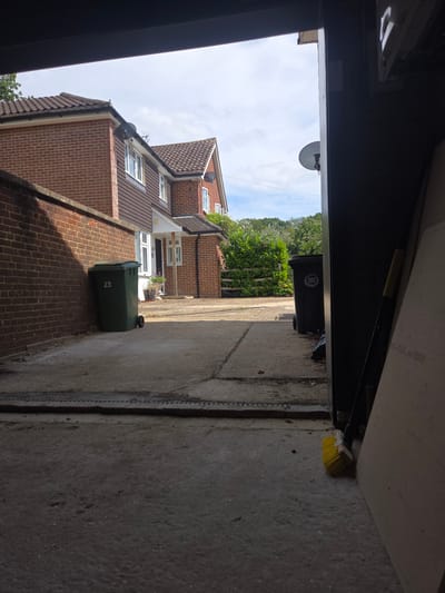 The image depicts an exterior view looking from a covered space, possibly a garage or porch, out towards a residential street on a cloudy day. The scene is oriented sideways, with the sky and houses appearing on the left and the ground and a wall on the right.
In the foreground, the edge of the covered structure is visible, with a yellow-bristled brush leaning against a concrete wall on the right side of the frame. Beyond this, a paved driveway or path leads towards a row of brick houses. A green wheelie bin, with the number "23" prominently displayed, sits on the path near the houses. Further down the street, another black wheelie bin is visible. The houses are made of red brick with tiled roofs and have multiple windows. Some houses have satellite dishes mounted on their roofs. Lush green trees and foliage are visible behind the houses, suggesting a suburban or semi-rural setting.
The sky is overcast, indicating a cloudy day, and the lighting suggests it is daytime. The overall impression is one of a typical residential street in the UK, likely Elmbridge given the location context. No people are visible in the image. The prominent text on the wheelie bin is "23."