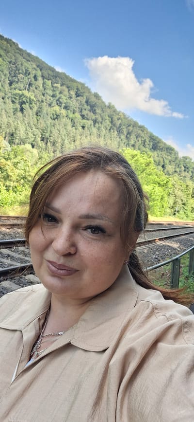 This media file is a portrait of a woman, taken outdoors. The woman is positioned in the foreground, looking towards the camera with a slight smile. She has brown hair and appears to be wearing makeup, including eyeliner and lipstick. Her clothing is a light-colored, collared shirt. A silver chain necklace is visible.
The background of the image shows a lush, green hillside covered in trees, suggesting a natural, possibly mountainous or hilly, environment. A bright blue sky with a few white clouds is visible above the hillside. To the right of the woman and extending into the background, there is a railway track with ballast and wooden sleepers, indicating the presence of a train line.
The lighting suggests it is daytime, likely sunny given the bright sky. The overall atmosphere appears relaxed and pleasant. There is no visible text in the image. The provided location context of "Maaracker, Germany" suggests the setting is in Germany.