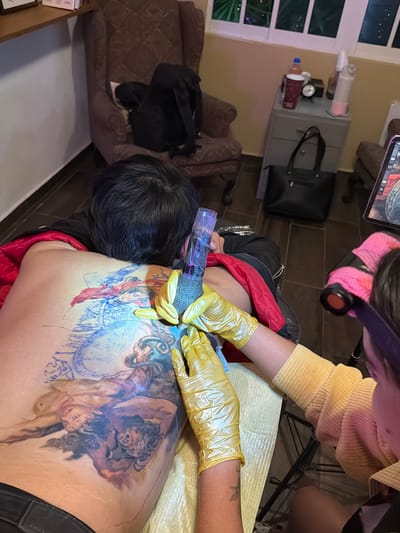 A person is receiving a tattoo on their back. The tattoo artist, wearing yellow gloves and a yellow corduroy shirt, is holding a tattoo machine. The tattoo design appears to depict figures in a classical style. The setting is an indoor space with a side table holding items such as a water bottle, a bag, and other various objects. There is no information about the location being in the city of Ciudad Madero, Mexico.
