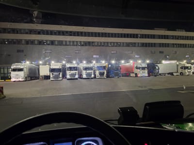 The image was taken at night from the interior of a truck cab, looking out at a row of parked semi-trailer trucks in Walldorf, Germany. The trucks are lined up in front of a large, modern building with multiple levels of windows. The building appears to be a warehouse or distribution center, as suggested by the loading docks visible at the rear of the trucks. The ground is paved with interlocking stones. The truck cabs are illuminated by various lights, including headlights, parking lights, and lights from the building. There are no people visible in the scene. The overall impression is of a busy logistics hub at rest during the night.