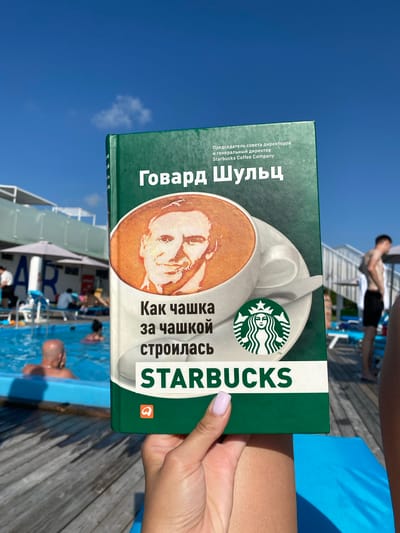 A woman's hand with pink nail polish holds a book with a green cover in front of a blurred outdoor swimming pool scene. The book's cover features a portrait of a man's face in a coffee cup, the Starbucks logo, and Russian text which translates to "Howard Schultz: How Cup by Cup Starbucks Was Built." In the background, several people are visible near or in the blue swimming pool. Umbrellas and lounge chairs are also present, suggesting a resort or public pool area. The sky is clear and blue. The location is not definitively Приамурский, Russia, as the image only provides visual information and no explicit location markers for that specific city.
