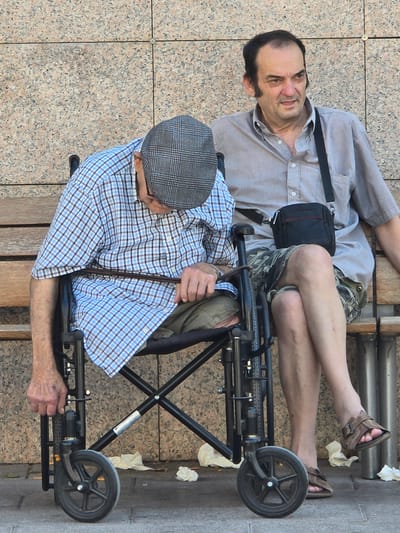 Two men are seated on a wooden bench in front of a granite wall. The man on the left is an elderly individual wearing a plaid shirt and a flat cap, seated in a wheelchair. He is leaning forward, with his head down and his right hand resting on the armrest of the wheelchair. The man on the right is younger, wearing a light-colored button-down shirt and camouflage shorts. He is looking to his left with a neutral expression. He has a small black bag slung across his body. Both men's legs are visible, and the younger man's legs are crossed. The ground in front of the bench has scattered white paper or tissue. This scene appears to be outdoors, likely in a public area, possibly Cornellà De Llobregat, Spain, given the information provided.