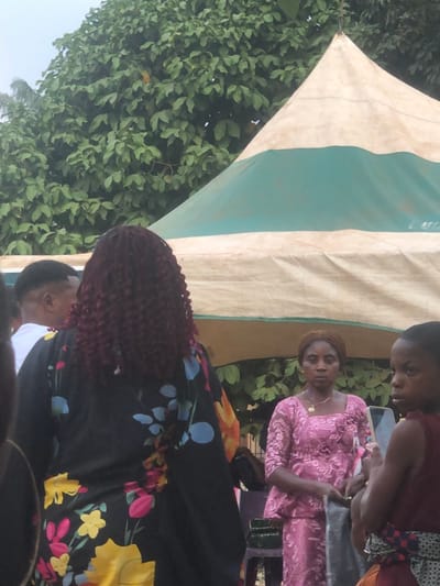 This image, captured in Awka Etiti, Nigeria, depicts an outdoor gathering under a large, cream-colored tent featuring a prominent green horizontal stripe. Lush green foliage is visible in the background, extending above and around the tent structure.

In the immediate foreground, a person with dark reddish-brown, tightly curled hair is seen from behind, wearing a loose-fitting black garment patterned with large, vibrantly colored yellow, blue, pink, and orange flowers. To their left, partially obscured, is the profile of an individual with a dark, short fade haircut, wearing a white shirt.

In the mid-ground, to the right of the person in floral print, a woman stands facing slightly towards the viewer's right. She has brown hair pulled back from her face and wears a pink lace two-piece outfit, consisting of a top and what appears to be a skirt. A gold necklace with a circular pendant is visible around her neck. Her left hand holds a dark, possibly fabric, object. To her right, a dark, low-lying object, possibly a chair or table, is partially visible. To the far right, a child with short dark hair and a reddish-brown top is partially visible, facing left. The people appear to be standing and observing the scene around them.
