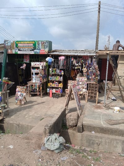 This image depicts a street-level commercial scene in Ijaiye, Nigeria. The prominent establishment on the left is "K.B. ENTERPRISES BUILDING MATERIALS," identified by a green and white sign displaying "IJAIYE, IBADAN." The shop features an open front with a corrugated metal roof, beneath which various items are displayed on shelves and tables. Merchandise includes numerous bottles of liquids (likely oils or chemicals), packaged small goods (possibly hardware or electrical components), and brooms. Two A-frame signs stand on the concrete path in front of the shop, one displaying text in red.
To the right, a shirtless male individual is positioned on a wooden scaffold or ladder structure, appearing to work on the upper section or roof of an adjacent building with a similar corrugated metal roof. A wooden utility pole with multiple overhead power lines stands between this individual and the shop. Another person is dimly visible inside K.B. Enterprises behind the merchandise displays.
The foreground consists of a dirt and gravel ground, with a large, light green bag secured with white string resting on it. This ground transitions to a concrete path directly in front of the shops, which is elevated and bounded by a concrete gutter or drainage channel. The sky above is partly cloudy.