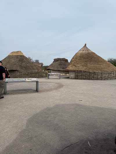 The image depicts a reconstructed ancient settlement, specifically three thatched roundhouses, located at the Stonehenge Visitor Centre near Amesbury, United Kingdom. The sky is uniformly overcast and light grey. The ground consists of a flat, light grey gravel-like surface.
In the foreground, a rectangular metal-framed information display panel is positioned, exhibiting text and images. A person, visible from the mid-torso down, stands to the left of this panel, wearing khaki trousers and a dark shirt, facing towards the panel. Behind this individual, a second person wearing a dark jacket is partially visible. Several indistinct figures are visible further back, near the leftmost roundhouse.
Three distinct roundhouses are present. The leftmost roundhouse features a rounded, conical thatched roof and a wattle fence forming its lower perimeter. The rightmost roundhouse has a steeper, more pointed conical thatched roof and a similar wattle fence. A third, smaller roundhouse with a thatched roof is situated in the mid-ground between the two larger structures. A second identical metal-framed information display panel is positioned centrally in the mid-ground, between the smaller roundhouse and the rightmost roundhouse. Green vegetation is visible beyond the structures in the background.
