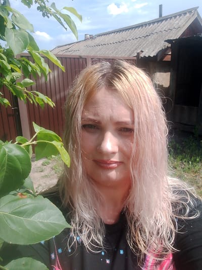 A medium shot photograph captures a Caucasian woman with blonde hair, exhibiting a slight smile and looking directly at the camera. She is positioned in what appears to be a rural or suburban outdoor setting. A large, green leaf is prominently in the foreground, partially obscuring the left side of the frame. Behind her, a brown wooden fence runs horizontally, and beyond that, a weathered, gray-shingled roof of a building is visible under a bright blue sky with scattered clouds. The overall environment suggests a sunny day.