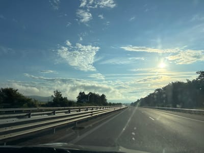 The image depicts a highway scene in Yazdach, Bulgaria, under a bright, partly cloudy sky. The sun is shining, creating a lens flare effect. The highway stretches into the distance, with a metal guardrail separating the lanes and the shoulder. Trees line the sides of the road, and in the background, hills are visible, partially obscured by clouds. The road surface appears to be asphalt, with white lane markings. The perspective suggests the photo was taken from inside a vehicle. There are no people or animals visible in the frame.