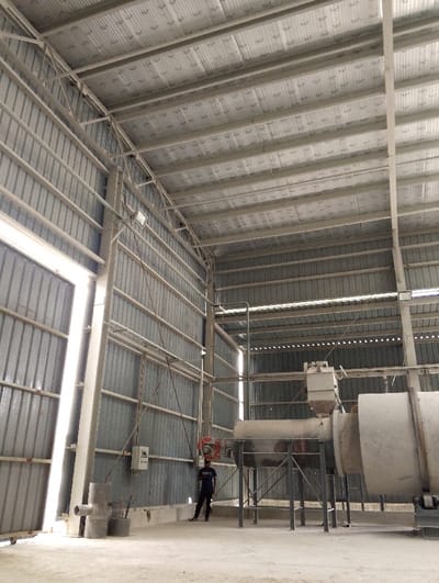 The image shows the interior of a large industrial building, likely a factory or warehouse. It is located in Pangasia, Bangladesh, at coordinates 22.694 latitude and 90.601 longitude. The building is constructed of metal, with corrugated metal walls and a corrugated metal roof.  There is a large industrial machine, possibly a cement mixer or grinder, in the center of the image. The machine is made of metal and is supported by a metal frame. A man in a blue shirt is standing near the machine, likely inspecting or operating it. There is a light fixture visible on the ceiling, and a metal door is located on the left side of the image. The floor is concrete. There are no other people in the image. The lighting is well-lit. There is no sign of activity or movement.  There is no evidence of any unusual or suspicious activity in the image.
