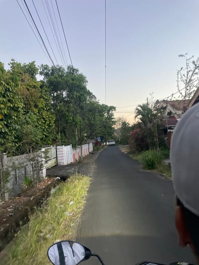 The image captures a perspective from a moving vehicle, likely a motorcycle, on a paved road in Manado, Indonesia. The road curves slightly to the left and recedes into the distance. On the left side of the road, there is a concrete wall adorned with red and white stripes, followed by lush green trees and foliage. Beyond the wall, glimpses of what appear to be houses can be seen. On the right side of the road, houses with weathered roofs and vegetation are visible. A white car is seen driving ahead on the road. Overhead, numerous power lines crisscross the sky. The sky is a pale blue, suggesting either early morning or late afternoon. The environment appears to be a residential area with a mix of natural greenery and man-made structures. The scene is dimly lit, with the headlight of the vehicle illuminating a portion of the road ahead.
