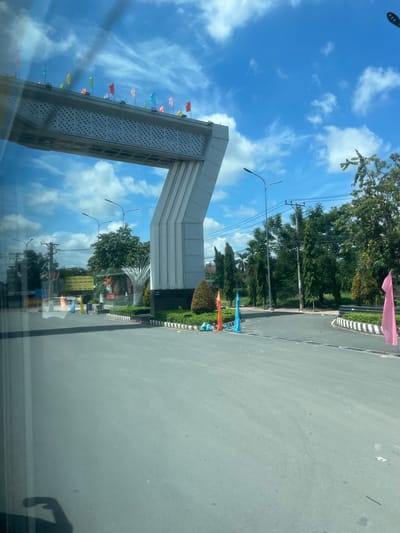 This is a vertically oriented, eye-level shot taken from inside a vehicle, looking out towards a bright, sunny day in Phường An Tịnh, Vietnam. The scene is dominated by a large, modern archway structure with a white, slatted surface and a slanted top. Numerous small, colorful flags in shades of red, yellow, blue, green, and pink are strung along the edge of the archway and on poles nearby, fluttering in the breeze.
Beyond the archway, a paved road curves to the right, bordered by a striped curb. Lush green trees and bushes line the roadside. Several streetlights are visible, and power lines crisscross the sky. The sky is a vibrant blue, dotted with fluffy white clouds, suggesting it is midday.
Reflections are prominent on the surface the photo is taken from, showing more of the sky, clouds, and the archway. A faint shadow of a person's arm and hand is visible in the lower right corner, indicating the photographer's presence.
In the background, a large, white metal sculpture with an abstract, fan-like design stands amidst the greenery. Behind this sculpture, several signs with Vietnamese text are visible, although the text is too small to be legible in the image. One sign has a yellow background with black text. The overall impression is one of a well-maintained public space or entrance, possibly to a town or a specific area, decorated for a festive occasion with the flags.