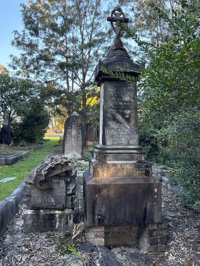 The image depicts a section of the Rookwood Necropolis cemetery in Chullora, Australia. In the foreground, a weathered stone tombstone stands prominently, adorned with a decorative cross featuring a rope motif at its apex. The tombstone exhibits inscribed text, partially obscured by a green leafy garland and the effects of time and weather. To the left of this central monument, a broken stone monument lies on the ground, its base crumbling and a section of stone chain detail visible.
The cemetery environment is characterized by overgrown grass, fallen leaves, and surrounding dense vegetation, including trees and bushes. Other gravestones and monuments, varying in size and design, are visible in the background, receding into the distance. The lighting suggests it is late afternoon or early morning, with long shadows cast by the trees and a clear, pale blue sky overhead. The overall impression is one of age and quiet solemnity, common to historical burial grounds.