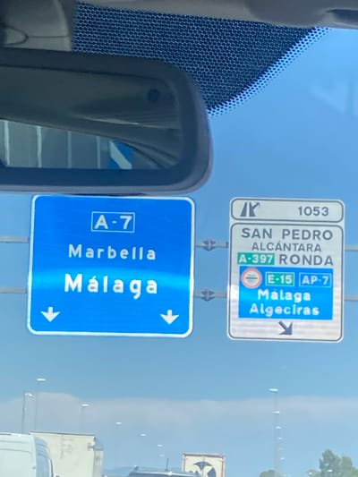 The image shows a road sign in San Pedro De Alcántara, Spain, on a bright, clear day. The sign is blue and white, with the text "A-7" at the top, followed by "Marbella" and "Málaga" below. Two white arrows point downwards on either side of the sign. To the right, another sign with a white background and black text indicates "SAN PEDRO ALCÁNTARA" and "A-397 RONDA". Below that, there are two smaller signs. One is circular and red, white, and black, and reads "E-15 AP-7". Below this, the text "Málaga Algeciras" is displayed with a downward-pointing arrow. In the background, vehicles can be seen on the road. The sky is a clear blue.