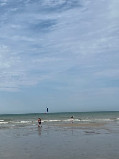 The image depicts a wide, sandy beach at Hautot-sur-Mer, France, under a bright, cloudy sky. The scene is dominated by the vast expanse of the ocean on the left and the wet sand reflecting the sky on the right, suggesting the tide has recently receded.

In the middle ground, two people are walking near the water's edge. They appear to be a couple, with one person in dark shorts and the other in a bikini. Further in the distance, another person is visible, and a kite, possibly for kitesurfing, is in the air. A second, smaller kite is also visible. The overall atmosphere is one of a pleasant day at the beach, likely during daylight hours. The weather appears to be warm and breezy, conducive to beach activities. The wet sand and the gentle waves indicate a calm sea. There is no visible text in the image.