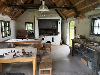This media file depicts the interior of a rustic workshop, likely at the historical-archaeological research and experience centre in Lejre, Denmark. The scene is a workshop focused on pottery or a similar craft, evidenced by the numerous small clay figures and pottery shards displayed on the tables.
The setting is characterized by its traditional architecture. The roof is thatched, supported by exposed wooden beams and rafters. The walls are plastered and appear somewhat aged, contributing to the historical atmosphere. The floor is made of concrete. Natural light enters through several windows, some with dark frames, and an open doorway in the background, suggesting it is daytime. The lighting within the workshop is a mix of natural light and industrial-style pendant lamps hanging from the ceiling.
Prominent objects include two large wooden worktables in the foreground and middle ground, laden with clay figures, tools, and pottery remnants. A large, traditional fireplace or kiln dominates one wall, with remnants of ash and char within its opening. Several wooden stools and benches are placed around the workshop, intended for seating. In the background, a dark wooden workbench is filled with more artifacts and tools. Various vessels, including large earthenware pots, are also visible. A fire extinguisher is mounted on a wall near the doorway.
The overall impression is that of a working studio, possibly a historical reenactment space, where visitors can engage in or observe traditional crafts. There are no people visible in the image. The mood is quiet and still, conveying a sense of industrious calm and historical immersion. There are no clear indicators of the weather outside, but the ample daylight suggests it is not dark. Visible text includes "HJÆLP OS HELP US" on two yellow signs affixed to the fireplace.