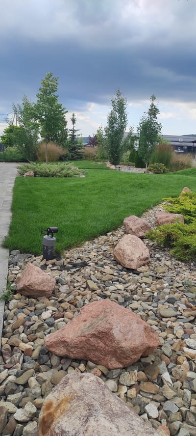 The media file displays a landscape scene at ground level, featuring a neatly mown green lawn bordering a decorative rock garden. The rock garden is composed of various sizes of stones, with several large, reddish-pink boulders interspersed among smaller, multi-colored pebbles. A dark, low-profile garden light is visible partially buried in the gravel, near the edge of the lawn.
Behind the lawn and rock garden, a line of trees and shrubs is visible. Some of the trees are slender with light-colored bark, while others are denser with green foliage. In the background, there is a building with some signage and what appears to be a paved walkway or driveway. The sky is overcast with dark, moody clouds, suggesting a late afternoon or evening on a potentially stormy day. The overall setting appears to be a landscaped outdoor area, possibly a park, garden, or commercial property. There is no clear indication of any people or specific activity taking place. The location context provided is Tulskiy, Russia.