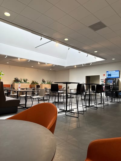 This image depicts a modern, unoccupied communal area or cafeteria located in Harderwijk, Netherlands. The space features a white grid panel ceiling with integrated recessed spotlights and track lighting, complemented by two large, arched skylights providing natural illumination. The walls are plain and light-colored. The floor consists of large, dark grey square tiles.
In the foreground, a round light grey table and the back of an orange upholstered chair are partially visible, with another orange chair partially visible in the bottom right. The midground and background contain multiple seating arrangements. These include rows of tall, square black tables with black metal bases, each paired with four black high-back bar stools. Adjacent to these are standard-height rectangular and square tables with light-colored tops and black metal bases, surrounded by chairs of varying designs, including light grey plastic chairs, darker grey chairs, and some with orange-backed seats. Along the left wall, brown upholstered booth seating is arranged with additional standard tables and chairs. Large wooden planters containing various green and purple leafy plants are strategically placed as decorative dividers.
In the background on the right, an open doorway leads to an interior room. A wall-mounted digital display screen exhibits information, including text (partially legible as "UPC") and an image of a vessel. Below the screen, a bank of at least three vending machines dispenses beverages and snacks. A red fire extinguisher is mounted on the wall near the vending machines. The area is well-lit, and no individuals are present; all furniture is unoccupied.