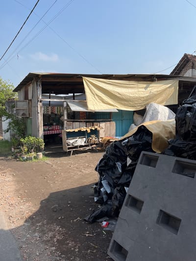 The image depicts a street scene in Gempolsari, Indonesia, featuring a small, open-air shop or warung. The structure is made of corrugated metal and wood, with a faded yellow tarp providing shade over the entrance. Inside, shelves are stocked with various packaged goods, and a person, partially obscured, appears to be tending to the shop. In front of the shop, a rustic wooden display stand holds items, including coils of rope and some white bags. To the right of the shop, a large pile of black plastic bags, some appearing to contain trash or recyclables, is stacked near a dark grey, rectangular object with recessed compartments, possibly a pallet or container. The ground is unpaved, with dirt, gravel, and scattered debris. The sky is clear blue with a few wispy clouds, and electrical wires are visible overhead. The scene is characterized by its simple, utilitarian construction and the presence of goods for sale and collected materials, suggesting a typical commercial area in a rural or semi-urban Indonesian setting.