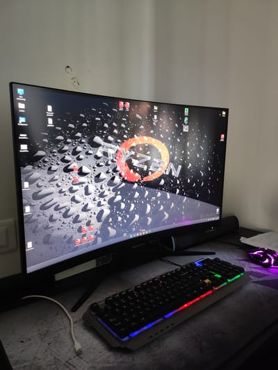 The image shows a computer setup with a large curved monitor displaying a wallpaper of water droplets and a stylized "RYZEN" logo. On the desk, a black keyboard with multi-colored backlighting is positioned in front of the monitor. A white USB cable is laid out on the desk, leading towards the left of the frame. A gaming mouse with purple lighting is visible on the right side of the desk, partially obscured by a document. The background consists of a plain light-colored wall with a few minor imperfections. The scene is indoors, likely in a home or office setting. There is no indication of the city of Grosseto, Italy, or any specific geographical location. The image focuses on the computer hardware and its immediate surroundings.