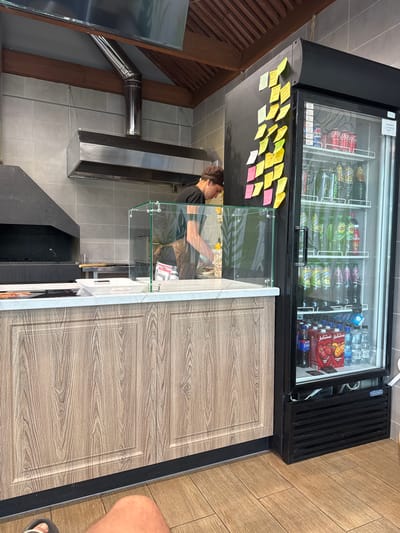 The image shows a food service establishment, possibly a pizza or fast food restaurant, in Baltasi, Russia. A person wearing a dark t-shirt and a dark cap is visible behind a counter, preparing food. The counter is made of a wood-like material with a marbled countertop. In front of the person, a glass partition separates them from the customer area.
To the right of the counter is a black commercial refrigerator filled with various beverages, including bottled water, soda cans (Coca-Cola), and juice boxes. The refrigerator has several shelves with drinks. Several colorful sticky notes are attached to the front of the refrigerator, likely containing orders or reminders.
Behind the counter, a stainless steel exhaust hood is installed above a cooking area that appears to be a pizza oven. The walls are tiled with light gray square tiles. The floor is made of wooden planks. The overall environment suggests a working kitchen or service area. The presence of a professional kitchen setup and commercial appliances indicates a business operating in Baltasi, Russia.