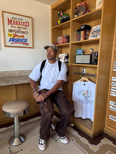 A man sits on a chrome and beige stool in front of a wooden shelf unit. He is wearing a white t-shirt, brown cargo pants, a camouflage baseball cap, and black and white high-top sneakers. He has tattoos on his left arm and is wearing silver rings and bracelets. On the shelves behind him are various items, including action figures, books, a framed picture, a speaker, and a white t-shirt hanging on a rack. To the left of the man is a framed poster with text that reads, "WHEN YOU'RE MARRIED, YOU'LL UNDERSTAND THE IMPORTANCE OF FRESH PRODUCE." The floor is covered in a mosaic tile pattern. The scene appears to be inside a deli or market, possibly in Marina del Rey, California.
